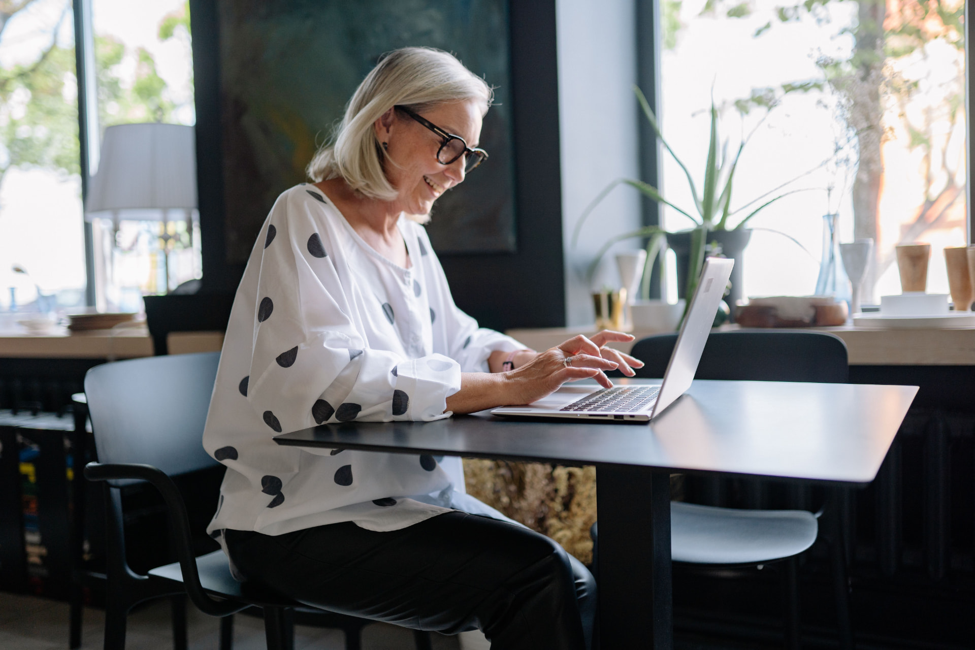 Smiling older woman with glasses working on a laptop at a bright cafe table